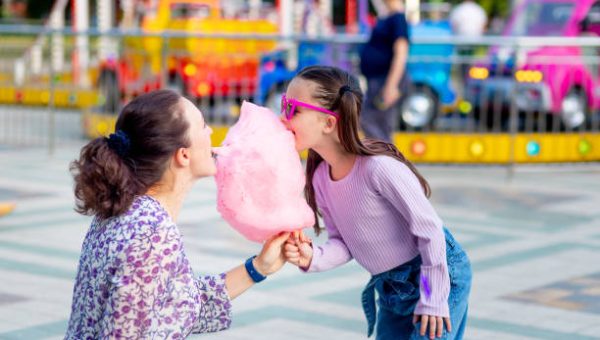 a child girl with her mother in an amusement park in the summer eating cotton candy and ice cream near the carousels, fooling around and laughing, the concept of family weekends and school holidays