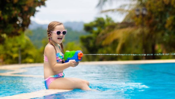 Child with water gun playing in swimming pool. Summer vacation with kids. Little girl jumping into water during exotic holiday in tropical island resort. Active outdoor sport for preschooler.